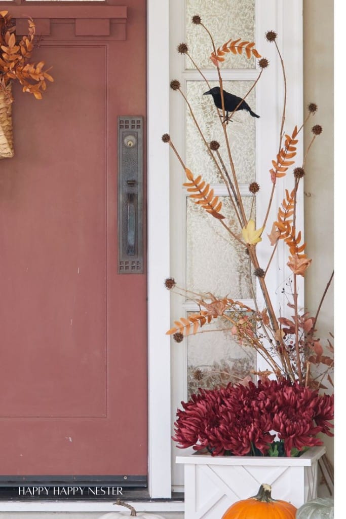 A fall-themed porch features a red door, a white pot with red flowers, and dried branches adorned with a black bird decoration. A small orange pumpkin rests nearby, creating a warm and festive autumn atmosphere.