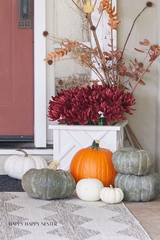 A fall-themed porch display with a white planter holding red flowers and orange leaves, surrounded by pumpkins in various sizes and colors, including orange, white, and green. A patterned gray rug is on the ground in front of a red door.