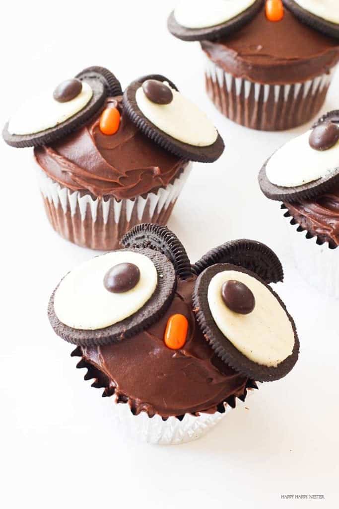 Four chocolate cupcakes decorated to look like owls make perfect Halloween treats for kids, with Oreo cookies and chocolate candies for eyes and orange candy for beaks, all set on a white surface.