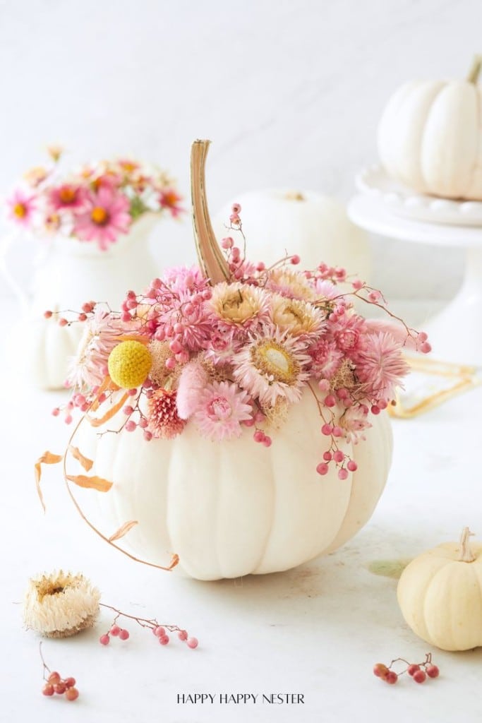 A white pumpkin is adorned with pink flowers and small berries, serving as a decorative centerpiece. In the blurred background, more floral arrangements featuring pumpkins and pink flowers can be seen.