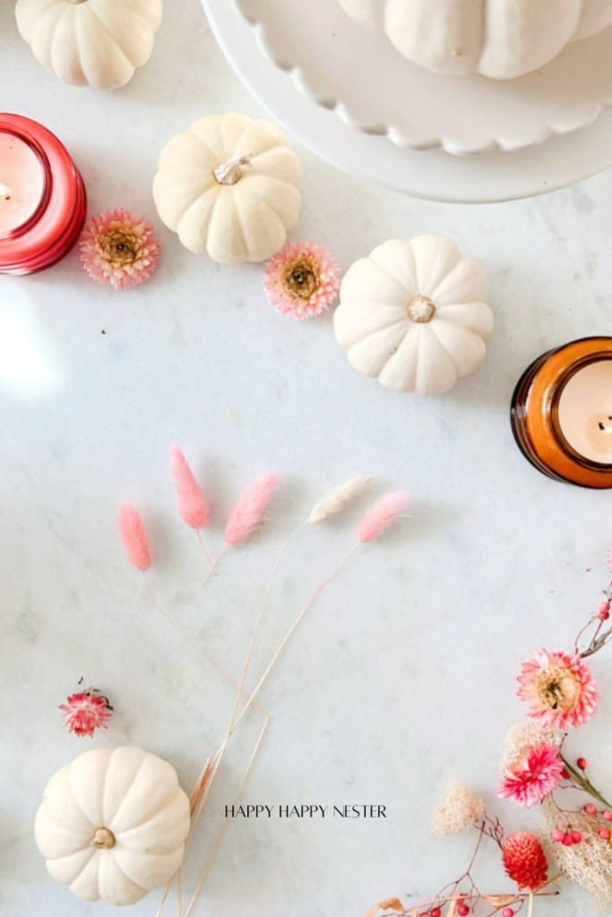 A flat lay of a fall-themed arrangement with small white pumpkins, pink and white flowers, and candles on a light surface.