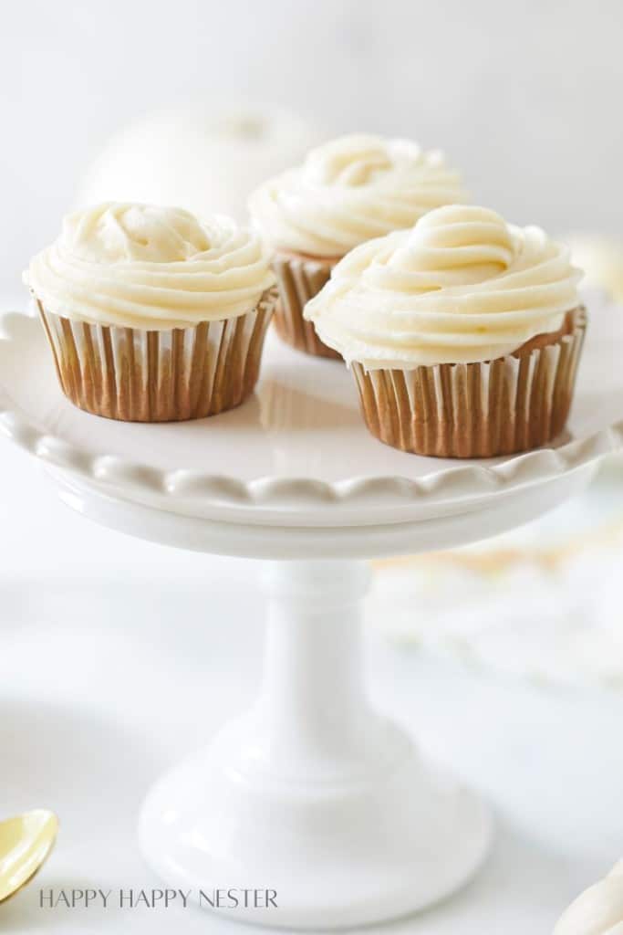 Three vanilla cupcakes topped with swirls of cream-colored frosting are elegantly arranged on a white cake stand, reflecting perfect fall dessert recipes. The softly blurred background beautifully emphasizes the cupcakes' presentation.