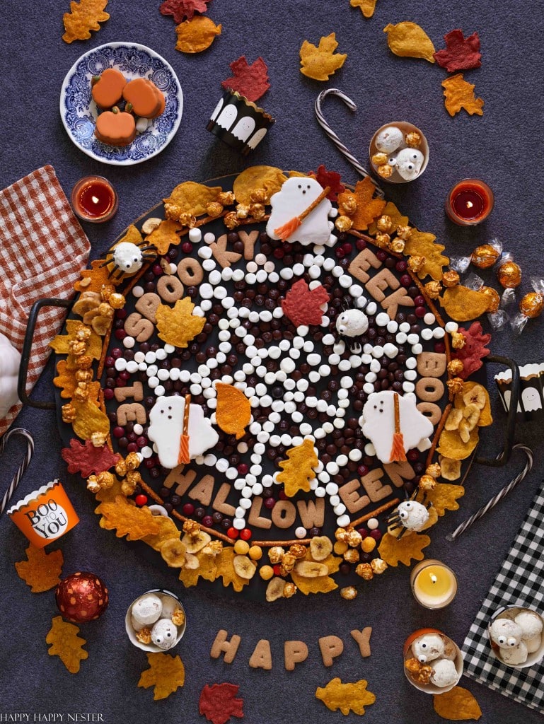 A festive Halloween-themed flat lay showcases a large tray of cookies in a spiderweb pattern, surrounded by candy, ghosts, and leaves. The words "Spooky" and "Eek" are displayed on a checkered cloth against a dark background—perfect halloween treats for kids.