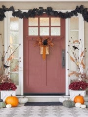 A welcoming fall-themed porch with a reddish-brown door decorated with a basket of autumn leaves. Black leaf garland frames the doorway. Pumpkins in various sizes and colors line the steps, complemented by potted plants on each side.