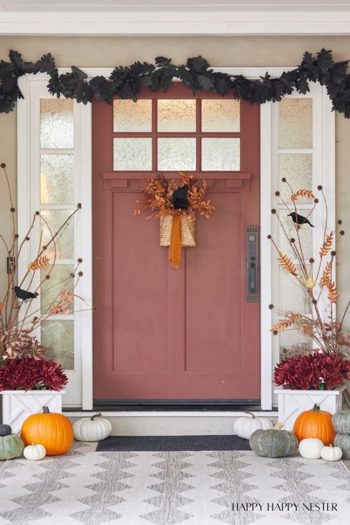 A welcoming fall-themed porch with a reddish-brown door decorated with a basket of autumn leaves. Black leaf garland frames the doorway. Pumpkins in various sizes and colors line the steps, complemented by potted plants on each side.