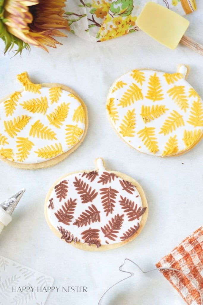 Three decorative sugar cookies shaped like pumpkins, each adorned with unique fern-like designs in yellow and brown icing, perfect for fall dessert recipes. A patterned cloth and baking tools are nearby on a light surface.