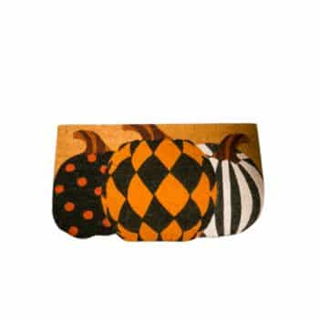 A decorative doormat featuring a Halloween theme with three pumpkins. One pumpkin is orange with black polka dots, the second is orange and black with a harlequin pattern, and the third is white with black stripes. The background is a solid orange color.