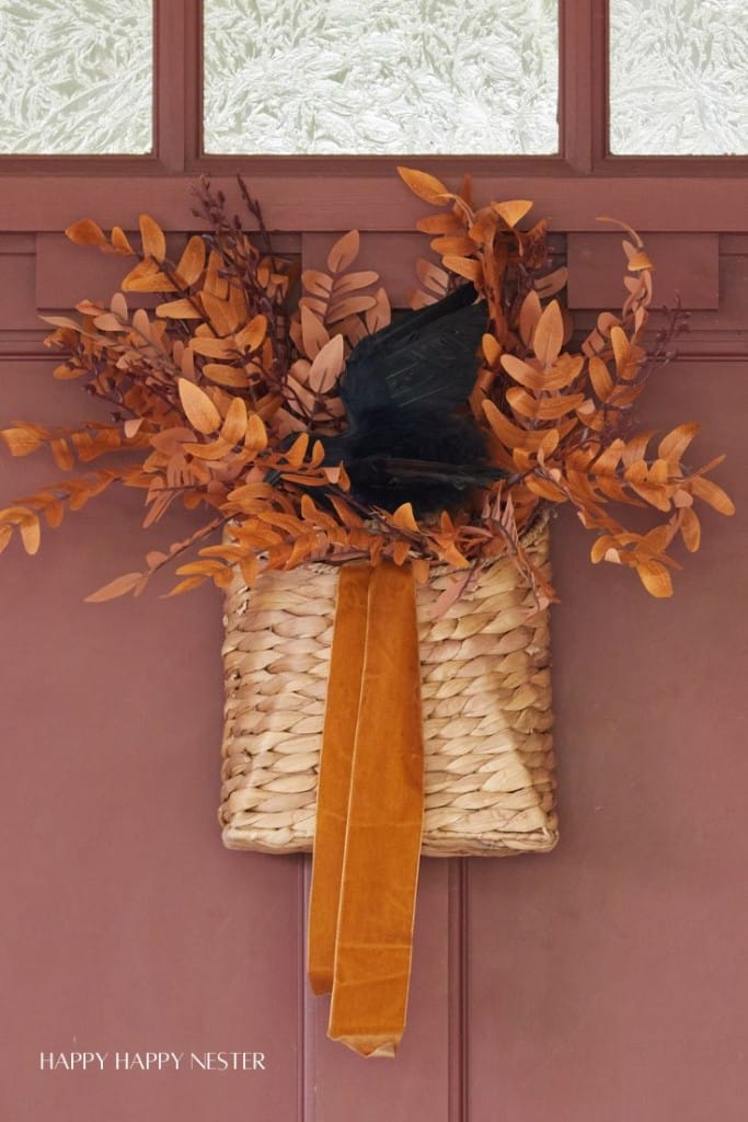 A cozy autumn-themed basket is hanging on a red door. It's filled with brown leaves and features a black witch's hat and an orange ribbon, capturing a festive fall atmosphere.