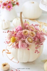 A white pumpkin is decorated with pink and beige dried flowers and berries on top. More white pumpkins and similar floral arrangements are in the background.