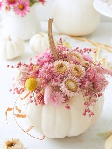 A white pumpkin is decorated with pink and beige dried flowers and berries on top. More white pumpkins and similar floral arrangements are in the background.