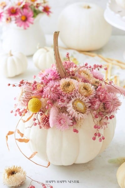 A white pumpkin is decorated with pink and beige dried flowers and berries on top. More white pumpkins and similar floral arrangements are in the background.