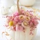 A white pumpkin is decorated with pink and beige dried flowers and berries on top. More white pumpkins and similar floral arrangements are in the background.