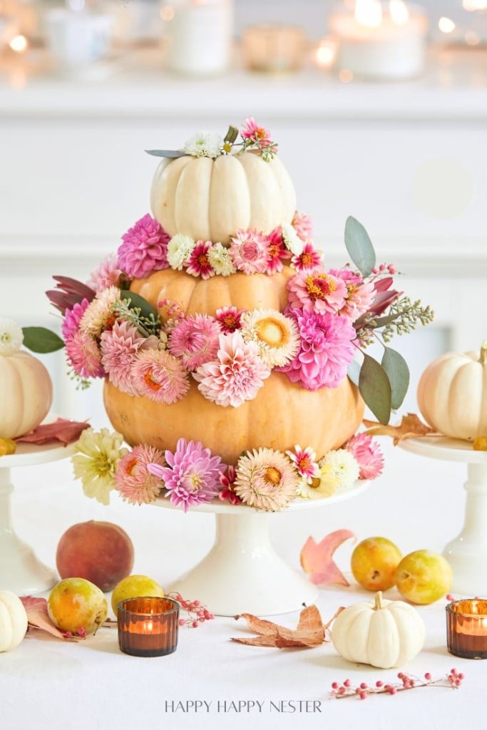 A tiered centerpiece made of stacked pumpkins, an easy pumpkin craft adorned with pink and white flowers, is surrounded by small pumpkins, candles, leaves, and fruit on a white table.