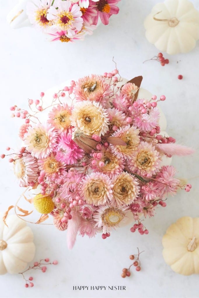 A bouquet of pink and cream dried flowers and berries is arranged on a white background, surrounded by small white pumpkins and more flowers. The scene has a soft, autumnal feel.