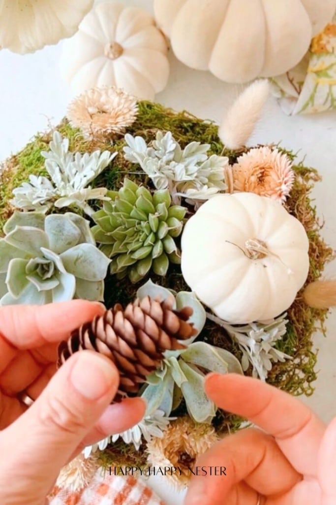 Hands carefully holding a small pinecone over a light autumn centerpiece, where DIY creativity shines with white succulent pumpkins, green succulents, and delicate flowers artfully arranged on a mossy base.