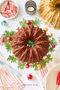 A beautifully decorated chocolate bundt cake sits on a glass cake stand with sprigs of greenery and colorful ornaments. The table features festive decorations, including a lit candle, candy canes, a gold bundt pan, and red and pink dishes—perfect for sharing your banana bread recipe using oil.