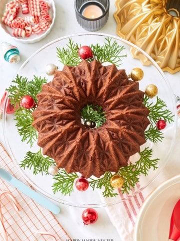 A beautifully decorated chocolate bundt cake sits on a glass cake stand with sprigs of greenery and colorful ornaments. The table features festive decorations, including a lit candle, candy canes, a gold bundt pan, and red and pink dishes—perfect for sharing your banana bread recipe using oil.