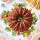 A beautifully decorated chocolate bundt cake sits on a glass cake stand with sprigs of greenery and colorful ornaments. The table features festive decorations, including a lit candle, candy canes, a gold bundt pan, and red and pink dishes—perfect for sharing your banana bread recipe using oil.