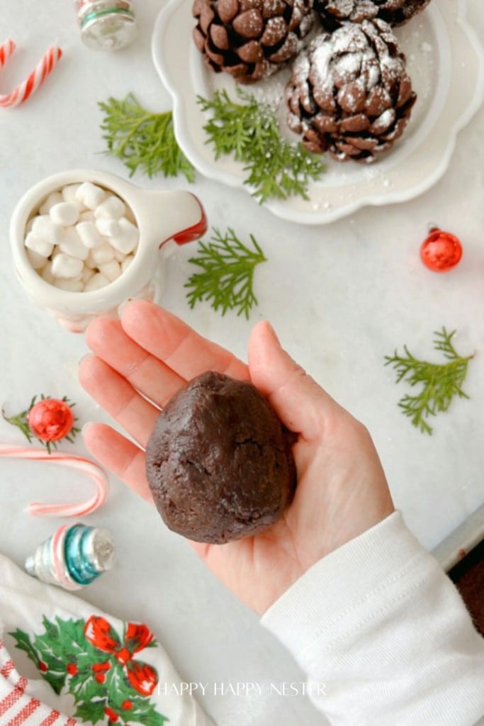 A hand holding a brownie dough ball over a table with holiday decorations. Nearby are a mug of hot chocolate with marshmallows, a plate of chocolate pastries dusted with powdered sugar, and festive ornaments.