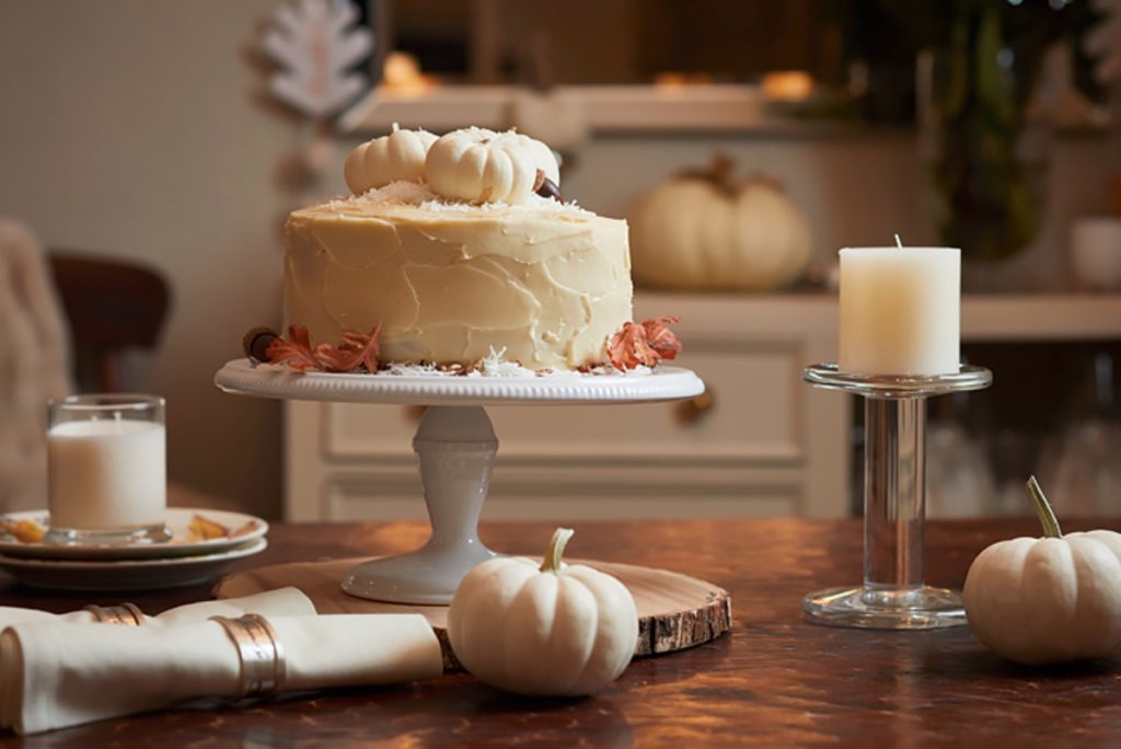 A decorated cake, reminiscent of fall pies and cakes, stands elegantly on a wooden table adorned with small white pumpkins. Nearby, white candles and additional pumpkins enhance the autumn-themed atmosphere.