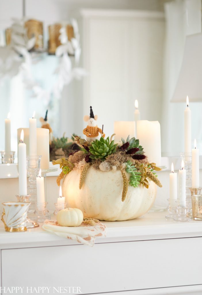 A white pumpkin, an example of easy pumpkin crafts, is adorned with succulents and autumnal accents, serving as a centerpiece on a white dresser. It's surrounded by lit candles and decorative items, creating a cozy and elegant atmosphere.