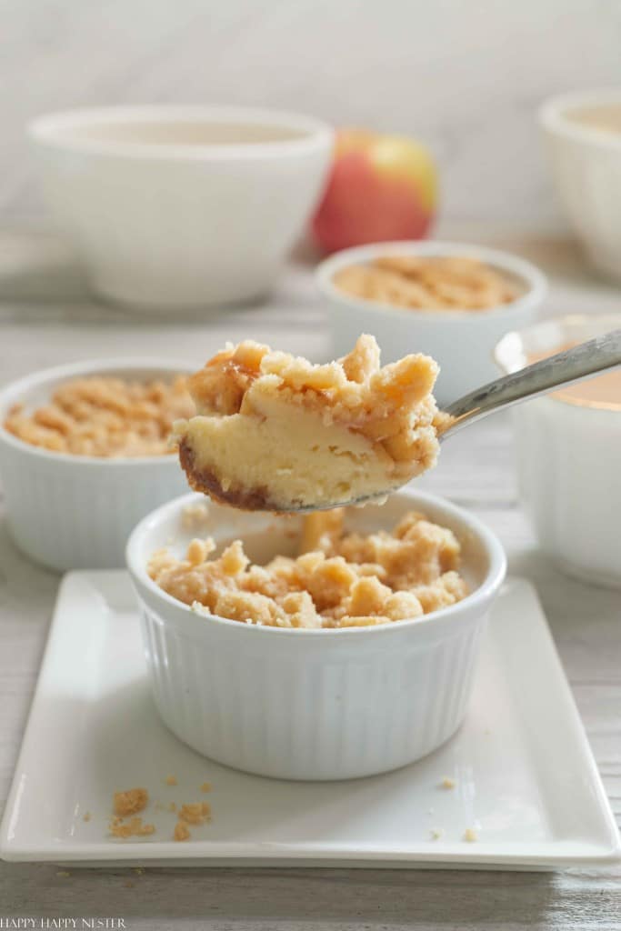 A spoon holding a scoop of creamy dessert with a crumbly top, reminiscent of fall pies and cakes, hovers above a white ramekin filled with the same treat. More ramekins with similar delights and a blurred apple in the background rest on a light wooden table.