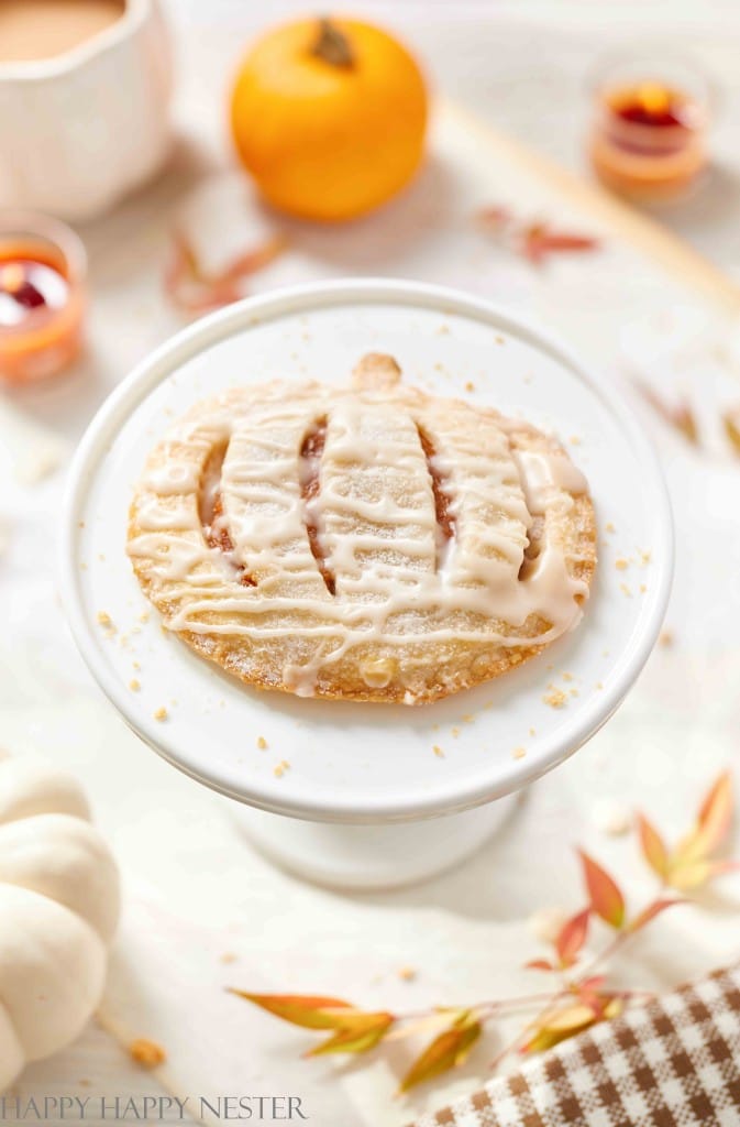 A pumpkin-shaped pastry with icing sits on a white stand, surrounded by small pumpkins and autumn leaves on a light-colored surface—a perfect centerpiece for Halloween treats for kids.