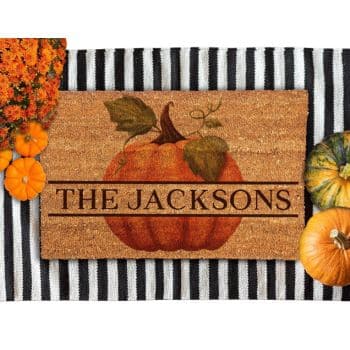 A doormat with "THE JACKSONS" written on it features a large pumpkin illustration. It's placed on a black and white striped surface. Surrounding the mat are small pumpkins and an orange flower arrangement.