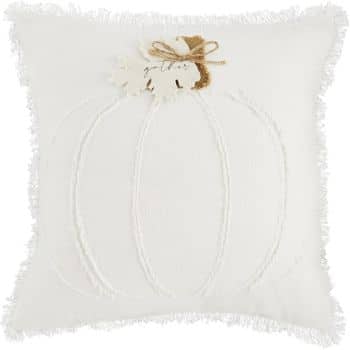 A white decorative pillow featuring a lightly embossed pumpkin design. Accents include brown leaves and a bow at the top. The pillow has fringed edges, giving it a cozy, autumnal look.