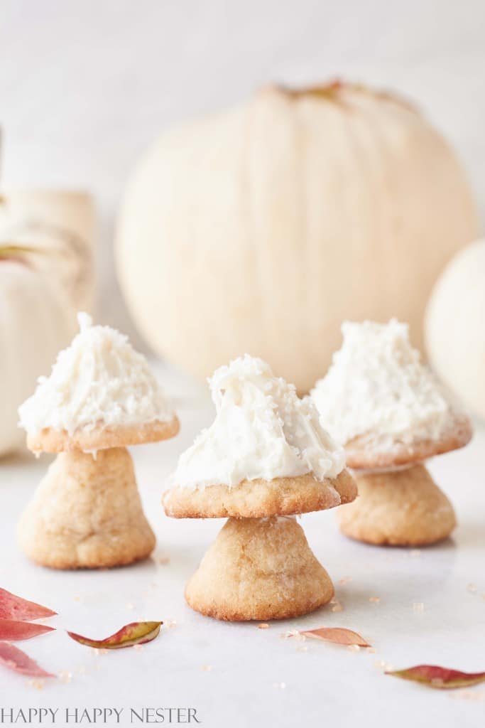 Three mushroom-shaped cookies coated with white frosting are arranged on a light surface. In the background, out-of-focus pumpkins add a seasonal touch. Scattered autumn leaves enhance the cozy fall atmosphere, making these Halloween treats perfect for kids.