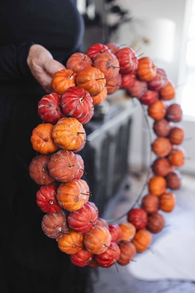 A person holds a festive wreath made of small pumpkins and adorned with autumnal flowers. This circular DIY creation is vibrant and perfect for seasonal decor, while the blurred background ensures all attention remains on the striking wreath.