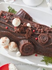 A chocolate yule log cake decorated with small meringue mushrooms and red berries sits on a white rectangular platter. Sprigs of evergreen are placed around the cake. The background features festive decorations and a red and white mug.