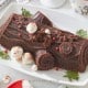 A chocolate yule log cake decorated with small meringue mushrooms and red berries sits on a white rectangular platter. Sprigs of evergreen are placed around the cake. The background features festive decorations and a red and white mug.