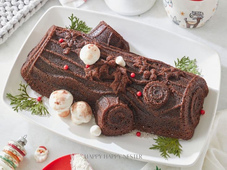 A chocolate yule log cake decorated with small meringue mushrooms and red berries sits on a white rectangular platter. Sprigs of evergreen are placed around the cake. The background features festive decorations and a red and white mug.