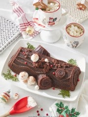 A chocolate Yule log cake, inspired by a Nordic Ware recipe, sits on a white platter adorned with red berries and green leaves. Nearby, festive mugs filled with marshmallows accompany a mini gingerbread house, all surrounded by holiday-themed linens and decor.