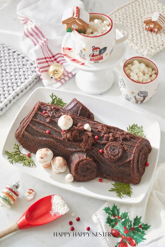 A chocolate Yule log cake, inspired by a Nordic Ware recipe, sits on a white platter adorned with red berries and green leaves. Nearby, festive mugs filled with marshmallows accompany a mini gingerbread house, all surrounded by holiday-themed linens and decor.