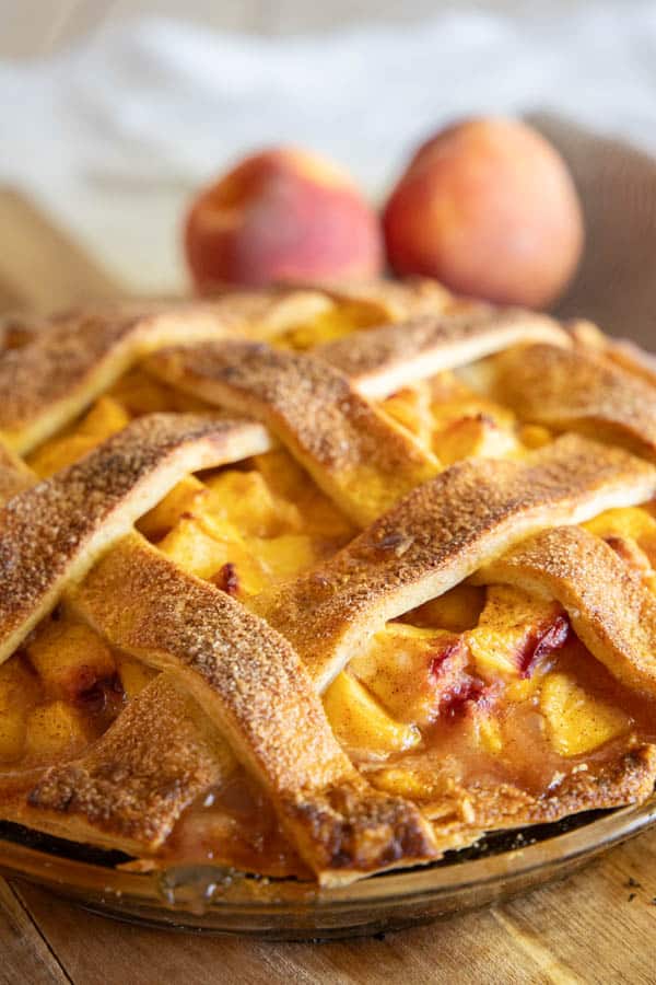 Close-up of a freshly baked peach pie with a golden brown lattice crust, set on a wooden surface. Two ripe peaches are in the background, echoing the rustic charm of your favorite Costco recipe, yet with a unique twist that delights any pie lover.