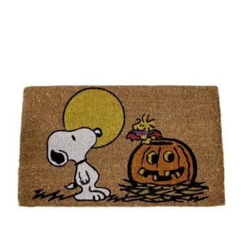 A doormat featuring Snoopy and Woodstock from the Peanuts comic strip. Snoopy stands howling at the moon, while Woodstock, dressed as a bat, sits on top of a carved jack-o'-lantern. The background is a simple tan color with a slight textured pattern.