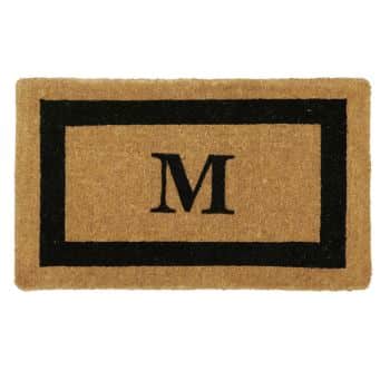 A rectangular doormat with a natural brown coir surface featuring a bold black letter "M" in the center. The letter is framed by a thick black border that runs parallel to the edges of the mat.