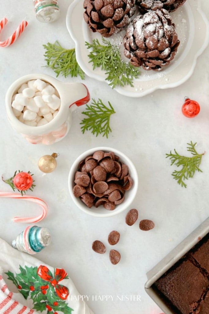 A festive holiday scene with pinecone brownies recipe dusted in powdered sugar on a white plate, alongside a cozy mug of marshmallows, a bowl of chocolate chips, evergreen sprigs, colorful ornaments, and a hint of pinecone brownies from your favorite recipe.