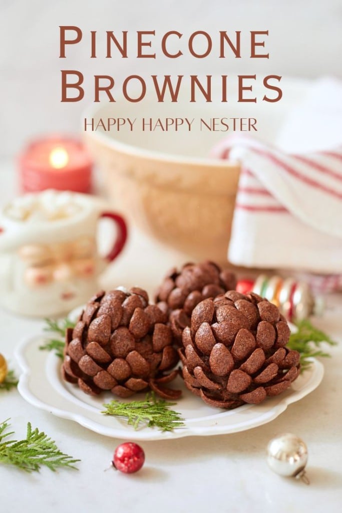 Three pinecone-shaped brownies sit on a white plate surrounded by festive holiday decor. In the background, a mug brims with marshmallows, folded dish towels add coziness, and a lit candle casts a warm glow. Text reads "Pinecone Brownies Happy Happy Nester.