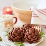 Two pinecone-shaped chocolate treats are on a white plate with evergreen sprigs. In the background, there's a striped napkin, a bowl, a candle, and a festive mug. Colorful ornaments are scattered around.