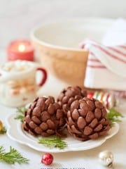 Two pinecone-shaped chocolate treats are on a white plate with evergreen sprigs. In the background, there's a striped napkin, a bowl, a candle, and a festive mug. Colorful ornaments are scattered around.