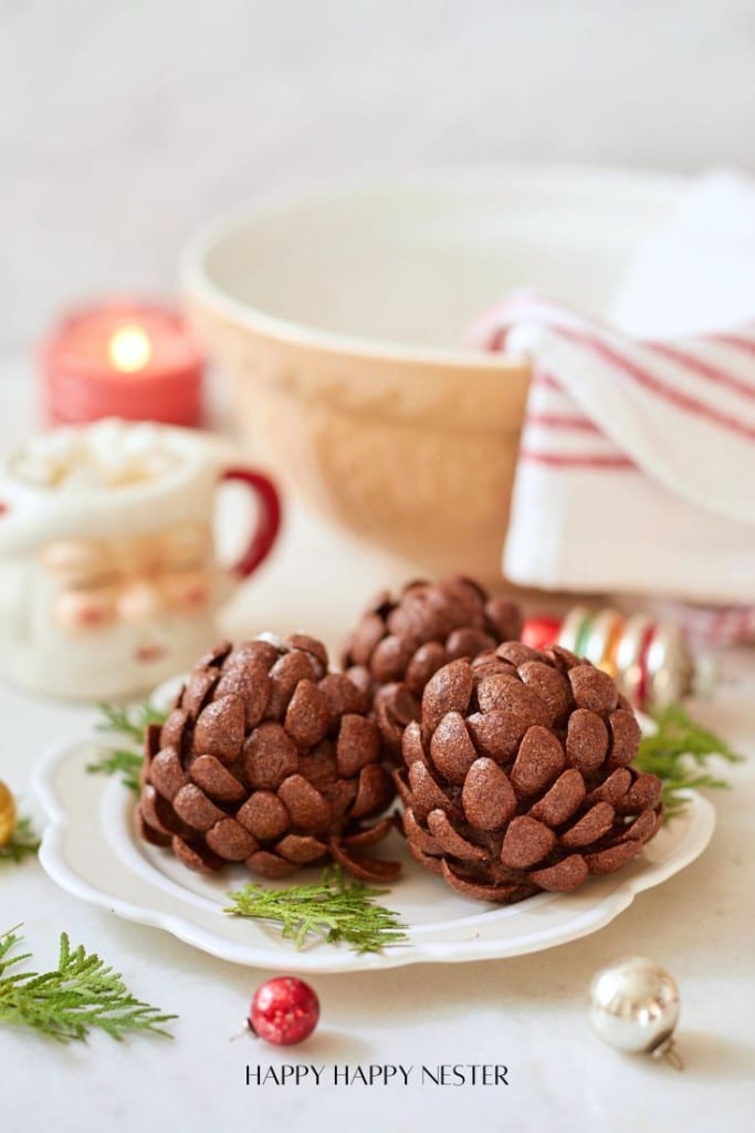 Two pinecone-shaped chocolate treats are on a white plate with evergreen sprigs. In the background, there's a striped napkin, a bowl, a candle, and a festive mug. Colorful ornaments are scattered around.