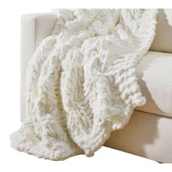 A plush, white knitted blanket draped over the arm of a cream-colored sofa. The blanket features a chunky, textured pattern, adding a cozy and inviting feel to the setting.