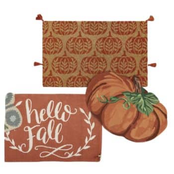 A decorative collection for fall includes a rectangular rug with a pattern of pumpkins, a doormat with the text "hello Fall" in white script on a rust background, and a pumpkin-shaped cushion with green leaves and a brown stem.