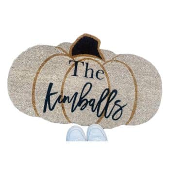 A pumpkin-shaped doormat with the text "The Kimballs" is displayed. The doormat is made of a woven material, and a pair of white shoes are partially visible at the bottom edge of the image.