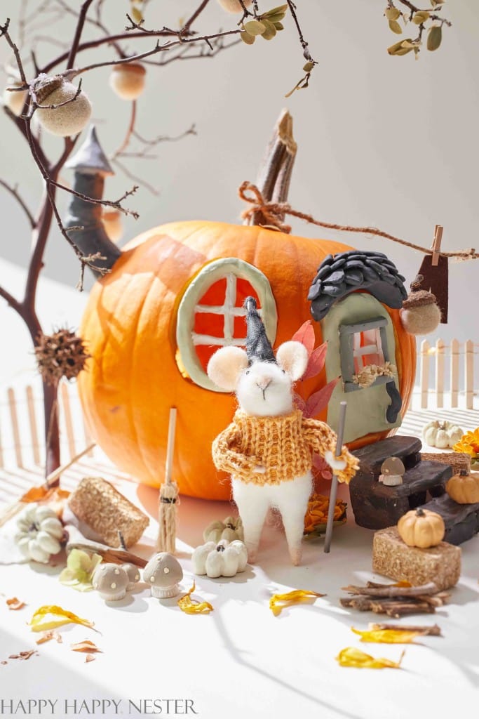 A whimsical scene features a pumpkin carved into a tiny house, showcasing easy pumpkin crafts with its windows and door. In front, a felt mouse in a knitted sweater stands amidst autumn leaves and miniature pumpkins, creating a cozy fall atmosphere.