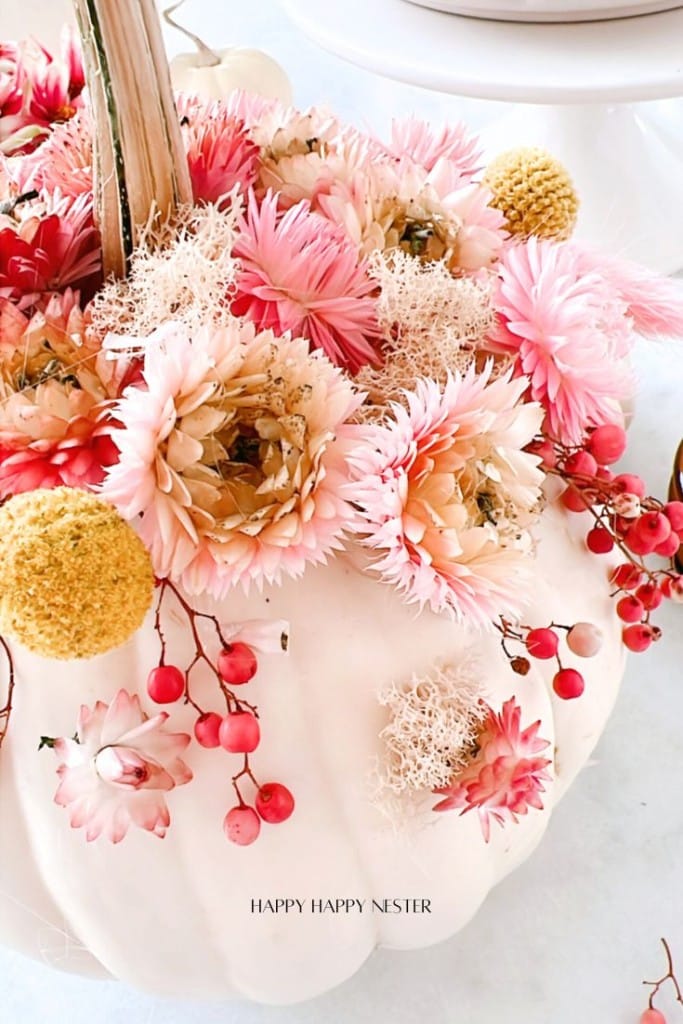 A white pumpkin decorated with pink and cream flowers, red berries, and tufts of white moss arranged artistically on top.