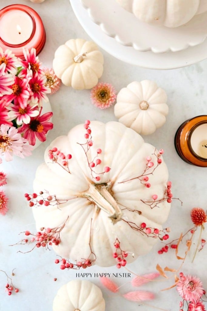 A festive fall arrangement featuring white pumpkins adorned with pink berries, surrounded by pink and red flowers, candles, and delicate leaves on a pale background.